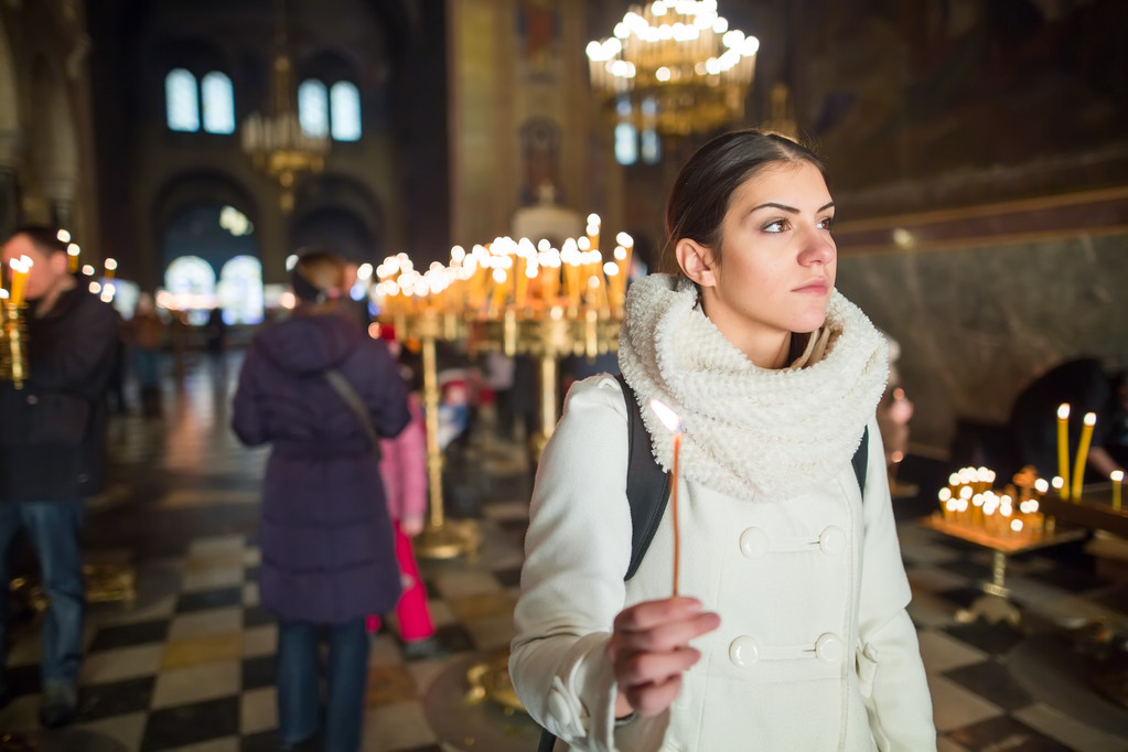 Young female lighting candles in a church during praying.Yellow votive candles burning.Woman praying to god at St. Alexander Nevsky Cathedral.Christianity - 写真・画像