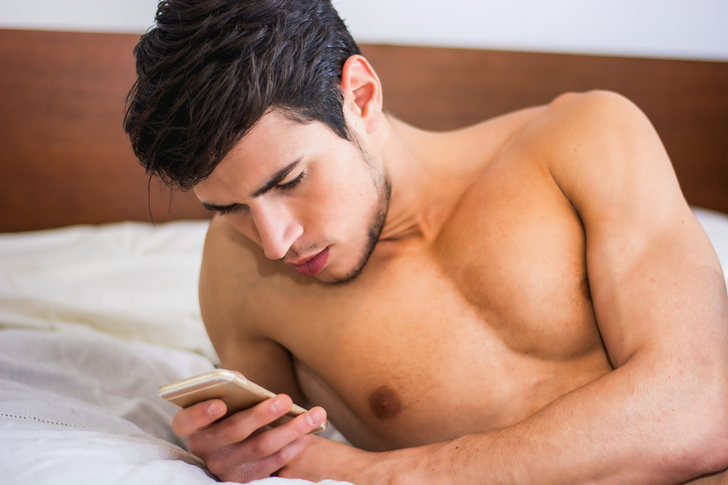 Όμορφος νεαρός στο κρεβάτι δακτυλογραφεί στο κινητό τηλέφωνο - Φωτογραφία, εικόνα