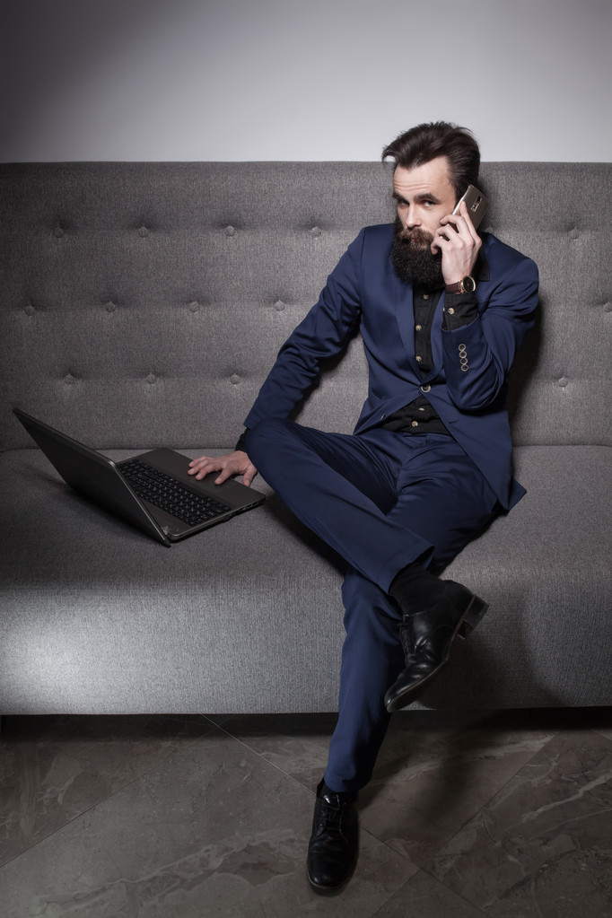 γενειοφόρος άνδρας ντυμένος με κοστούμι και με φορητό υπολογιστή και να μιλήσουμε στο τηλέφωνο; - Φωτογραφία, εικόνα