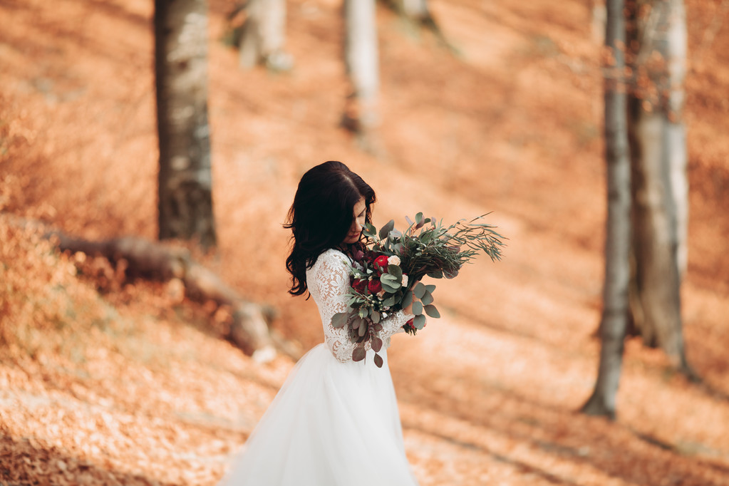 Superbe mariée brune en robe élégante tenant bouquet posant près de la forêt et du lac
 - Photo, image