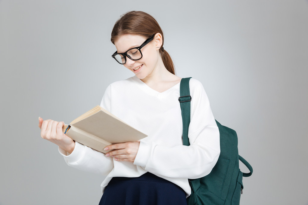 Cheerful lovely teenage girl in glasses with backpack reading book  - Photo, Image