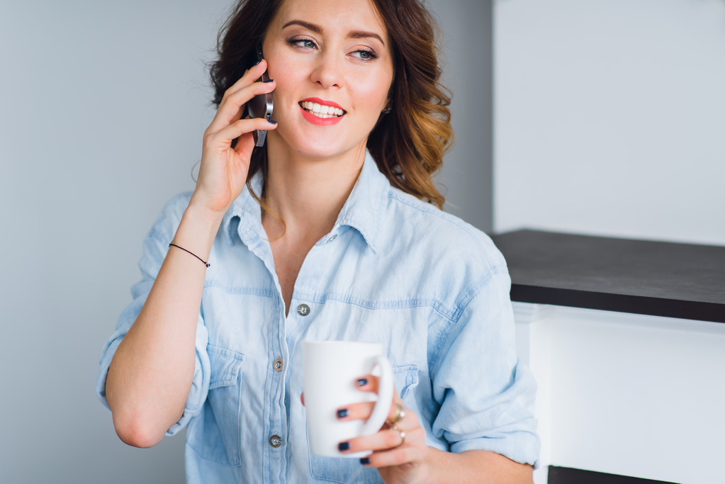 Красивая улыбающаяся женщина с кофейной чашкой разговаривает по сотовому телефону дома
. - Фото, изображение