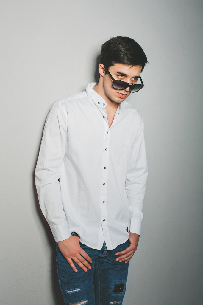 Jovem de shorts e camisa branca está sorrindo de pé perto da parede com óculos
 - Foto, Imagem
