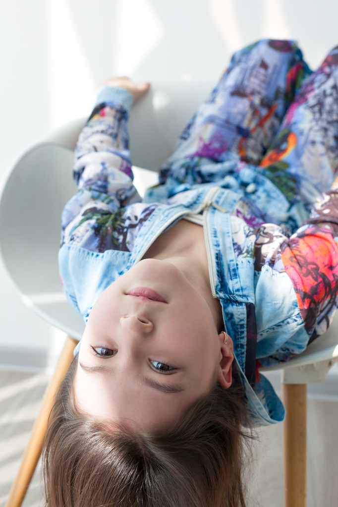 Ritratto di una bambina di moda su una sedia bianca. Elegante bambina con i capelli lunghi si siede a testa in giù. Foto modello bambino cool alla moda
. - Foto, immagini