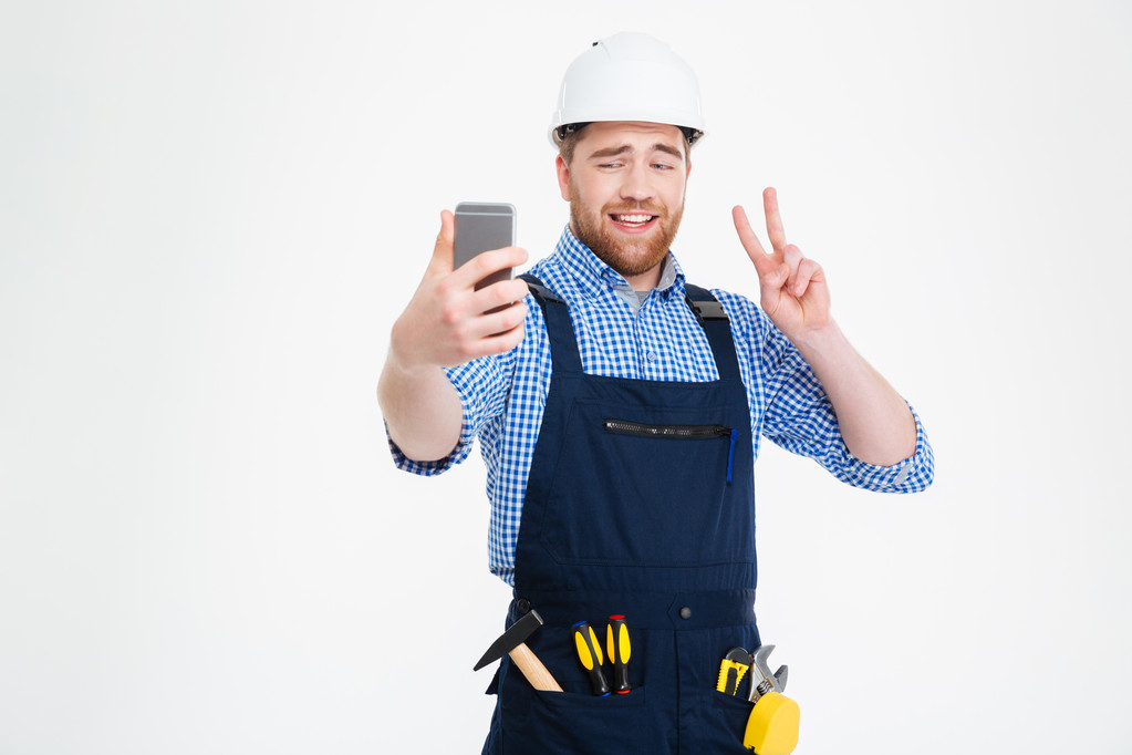 Усміхнений будівельник показує знак миру і робить селфі зі смартфоном
 - Фото, зображення