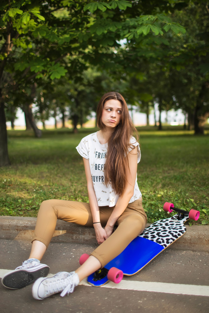 Młoda dziewczyna z smutną twarz siedzi w parku z longboard. Na zewnątrz, styl życia - Zdjęcie, obraz