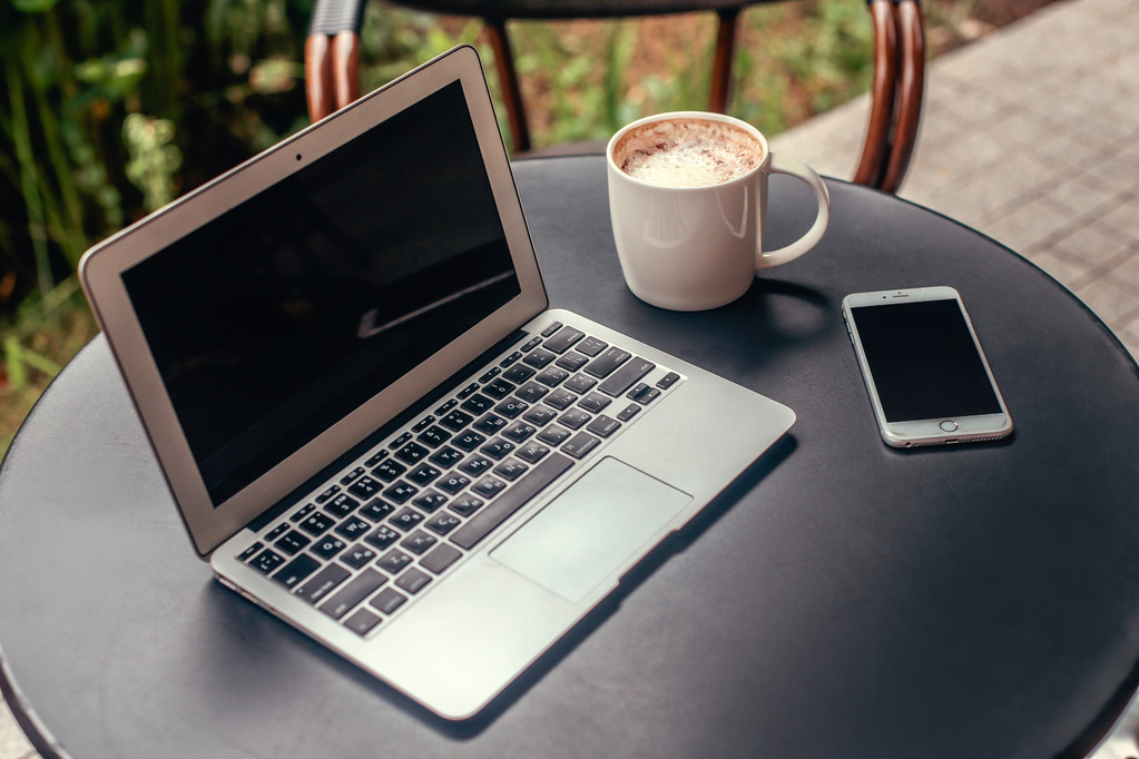 Kannettava tietokone, älypuhelin ja kuppi kahvia pyöreä musta pöytä kahvilassa
 - Valokuva, kuva