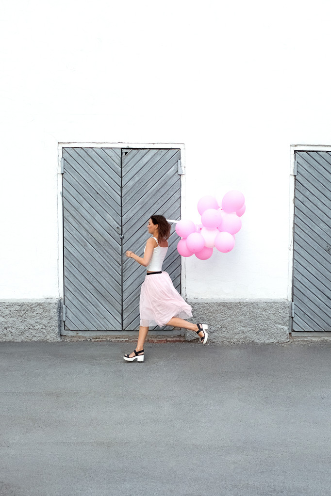 Vista laterale della giovane bruna che corre con palloncini rosa lungo le porte in legno per la strada.Copia spazio
 - Foto, immagini