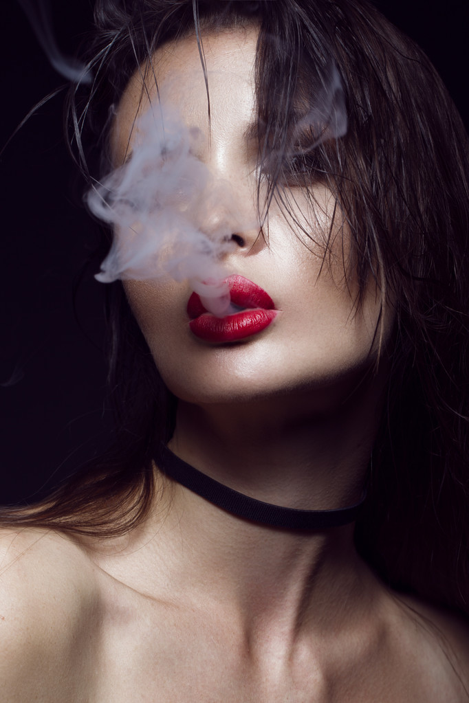 Belle fille brune sexy avec un maquillage lumineux, lèvres rouges, fumée de la bouche. beauté visage
. - Photo, image