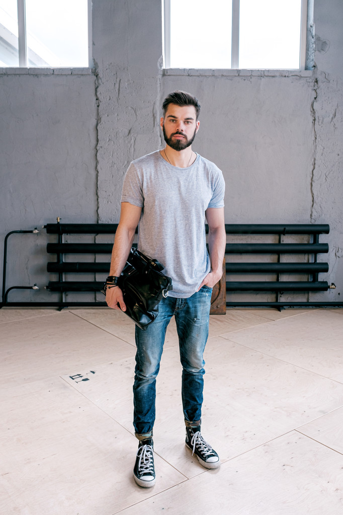 Portrait of handsome bearded stylish man holding handbag and looking at camera.Old wall with heating system and window on background. - Photo, Image