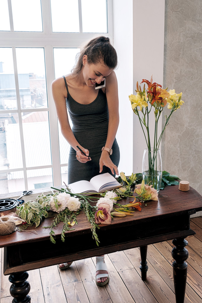Retrato de florista feliz y ocupado hablando por teléfono y escribiendo en cuaderno en la mesa con flores
 - Foto, imagen