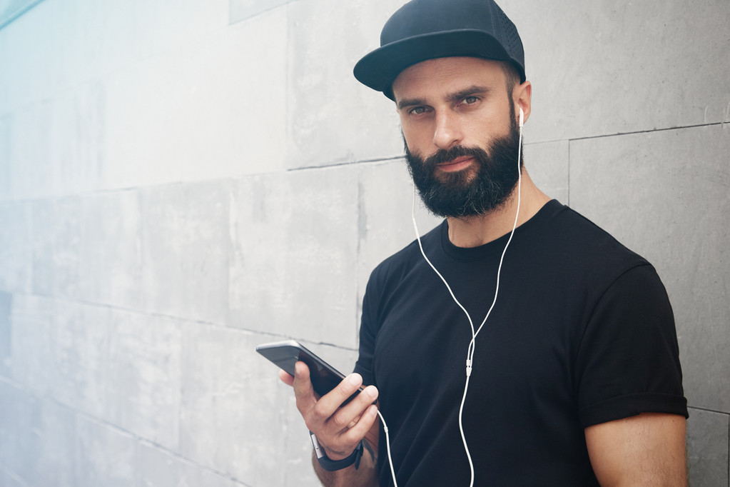 Bearded Muscular Man Wearing Black Tshirt Blank Snapback Cap Summer Time.Young Men Smiling Opposite Empty Gray Concrete Wall Background Using Smartphone Headphones.Horizontal Mockup.Side View. - Photo, Image