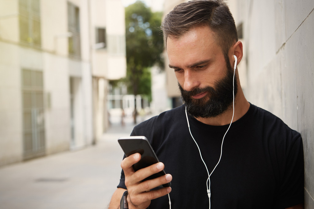 Бородатий м'язистий чоловік у чорній футболці Бланк Snapback Cap Літній час. Молоді чоловіки використовують смартфон навушники Дивлячись екран Усміхаючись. Горизонтальна
. - Фото, зображення