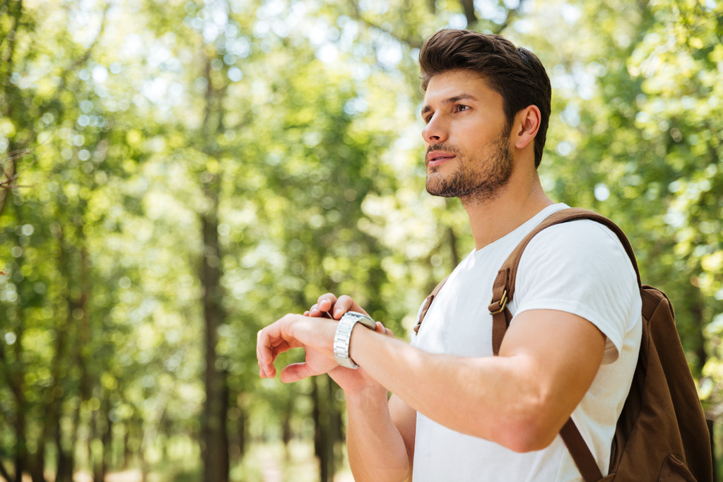 Nachdenklicher junger Mann mit Rucksack blickt auf Uhr im Wald - Foto, Bild