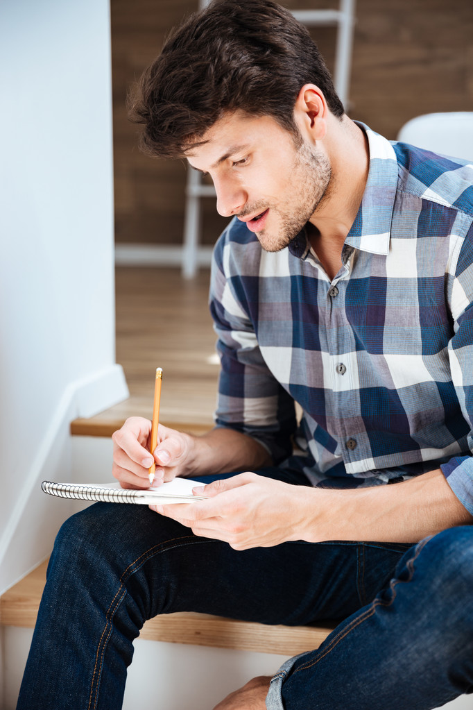 Keskittynyt nuori mies istuu ja kirjoittaa muistilehtiössä kotona
 - Valokuva, kuva