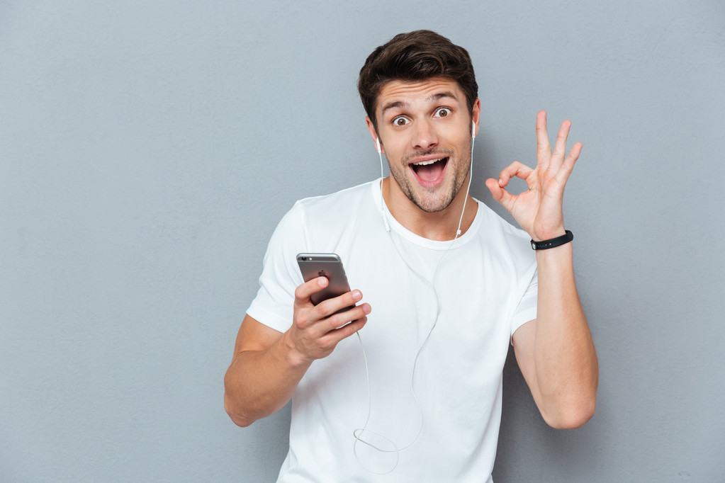 Cheerful man with earphones holding smartphone and showing okay sign - Photo, Image