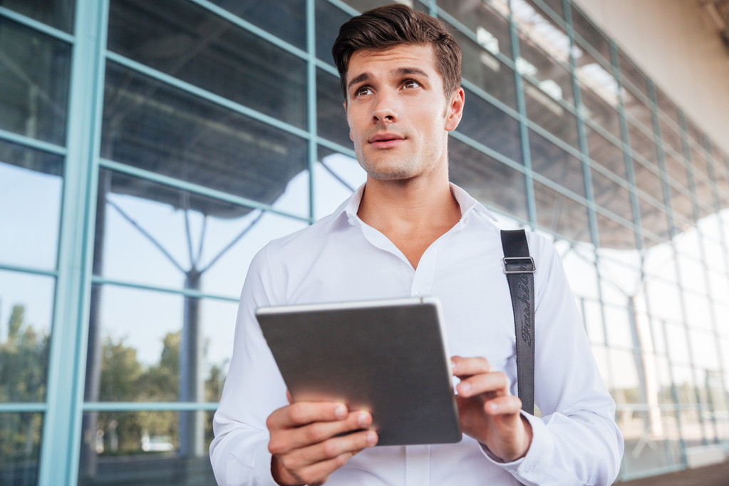 Συλλογισμένος όμορφος νεαρός επιχειρηματίας στέκεται και χρησιμοποιώντας tablet - Φωτογραφία, εικόνα