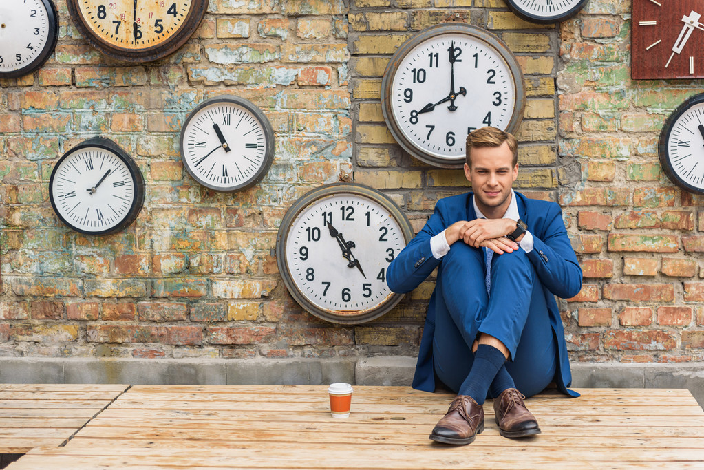 Homem de terno sentado perto da parede com relógios
 - Foto, Imagem