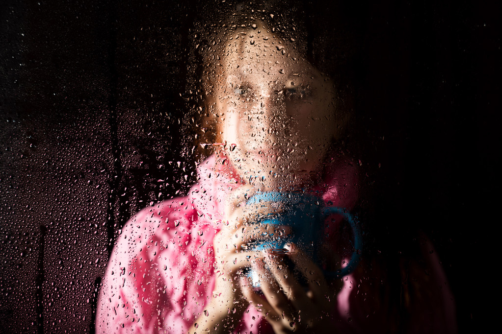 Portret van de jonge triest vrouw achter het raam in de regen met regen druppels op het. meisje met een kopje warme drank - Foto, afbeelding