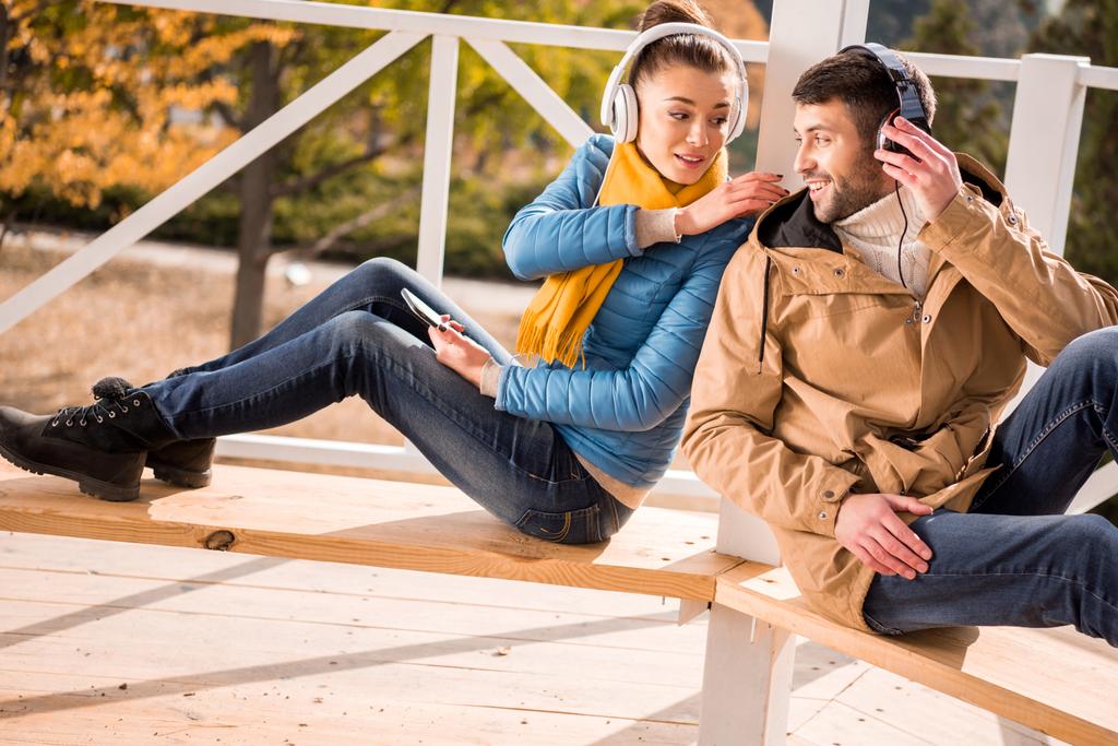 Νεαρό ζευγάρι κάθεται σε παγκάκι στα ακουστικά - Φωτογραφία, εικόνα