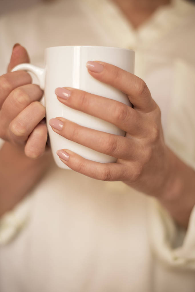 Girl is holding white cup in hands. White mug for woman, gift. Female hands holding hot cup of coffee or tea in morning. Selective focus - Photo, Image