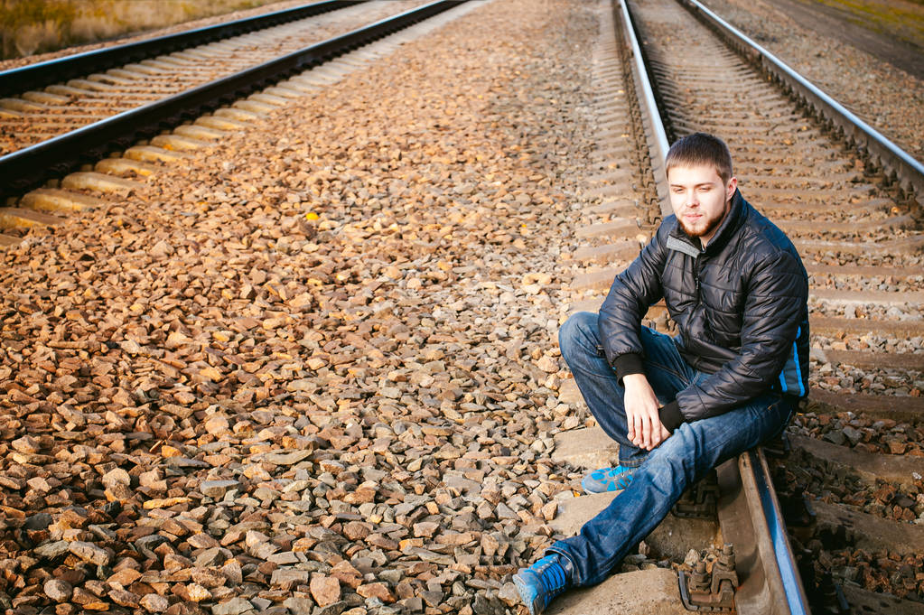 若い男は、トラックの上に座って。ジャケットとジーンズ、列車が到着するまでのレールとレールの枕木の上に座って男のひげを生やした。秋の風景と砂利の背景に、新しい旅行を見込んでいます。 - 写真・画像