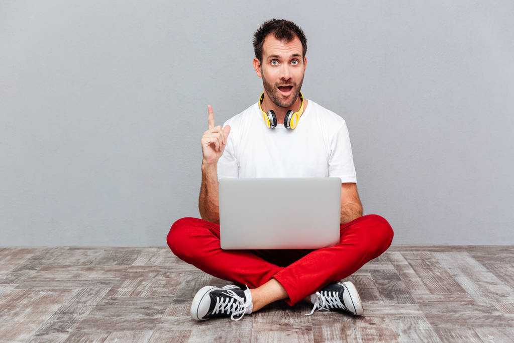 Мужчина сидит на полу с ноутбуком и показывает пальцем вверх
 - Фото, изображение