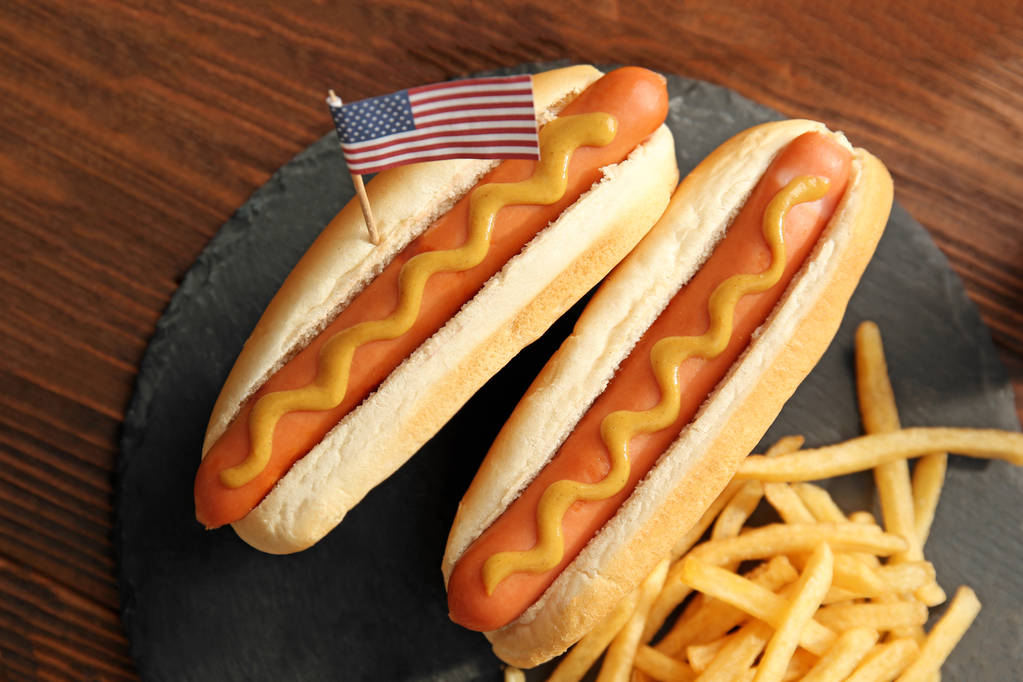 Perros calientes con mostaza y pequeña bandera de Estados Unidos en placa de pizarra
 - Foto, Imagen