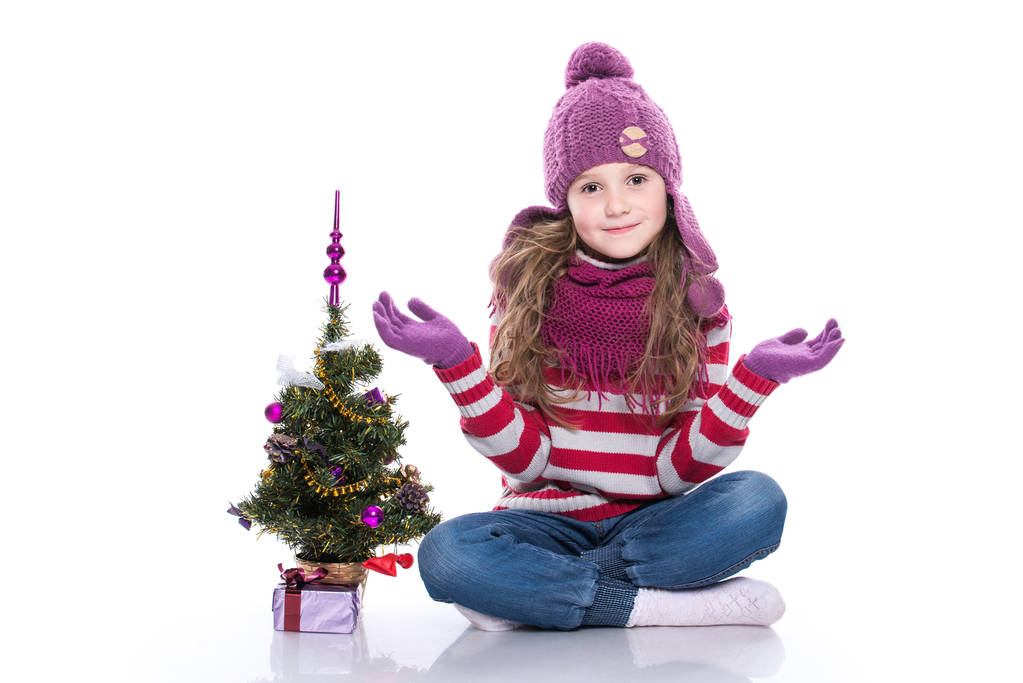 Mor örgü atkı ve şapka, Noel ağacı ve hediye beyaz arka plan üzerinde izole yakınında oturan sevimli gülümseyen küçük kız. Kış giysileri ve Noel kavramı. - Fotoğraf, Görsel
