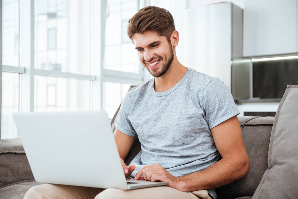 Szczęśliwy młody człowiek siedzi na kanapie w domu z laptopem - Zdjęcie, obraz