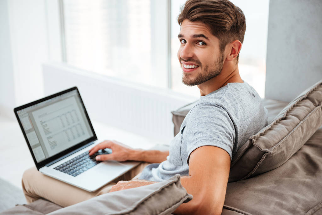 Szczęśliwy młody mężczyzna siedzi z laptopa i patrząc na kamery - Zdjęcie, obraz