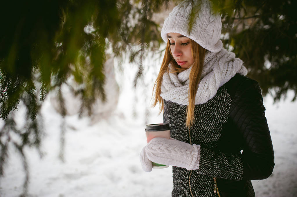 Портрет милой женщины в белом шарфе и шапке вязаном пальто на открытом воздухе на фоне снега и размытых еловых ветвей зимой
 - Фото, изображение