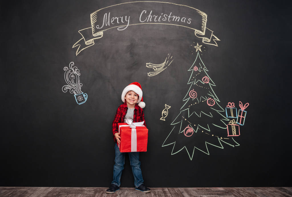 Criança bonito segurando grande presente perto de desenho de Natal no quadro negro
 - Foto, Imagem