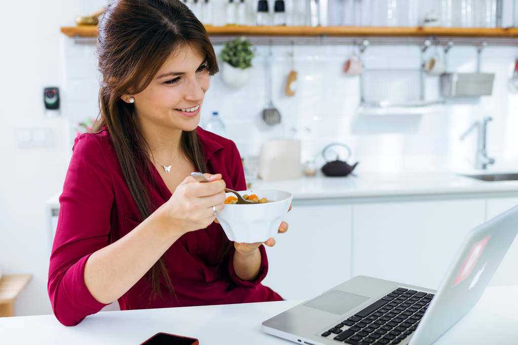 schöne junge Frau arbeitet mit Laptop beim Essen in der Küche. - Foto, Bild