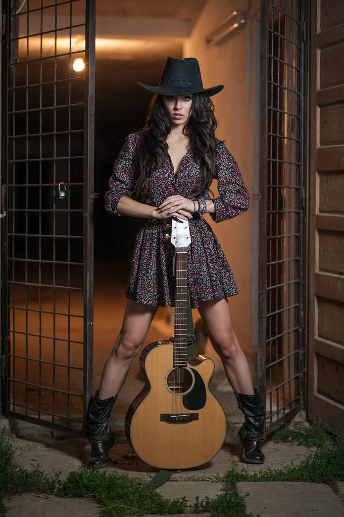 Ελκυστική γυναίκα με χώρα κοιτάξτε, γυρίστηκε σε εσωτερικούς χώρους, αμερικανική χώρα στυλ. Κορίτσι με μαύρο καπέλο και κιθάρα. Όμορφη μελαχρινή με κοντό φόρεμα και μπότες ποζάρει μόδας σε εσωτερικούς χώρους με μια κιθάρα - Φωτογραφία, εικόνα
