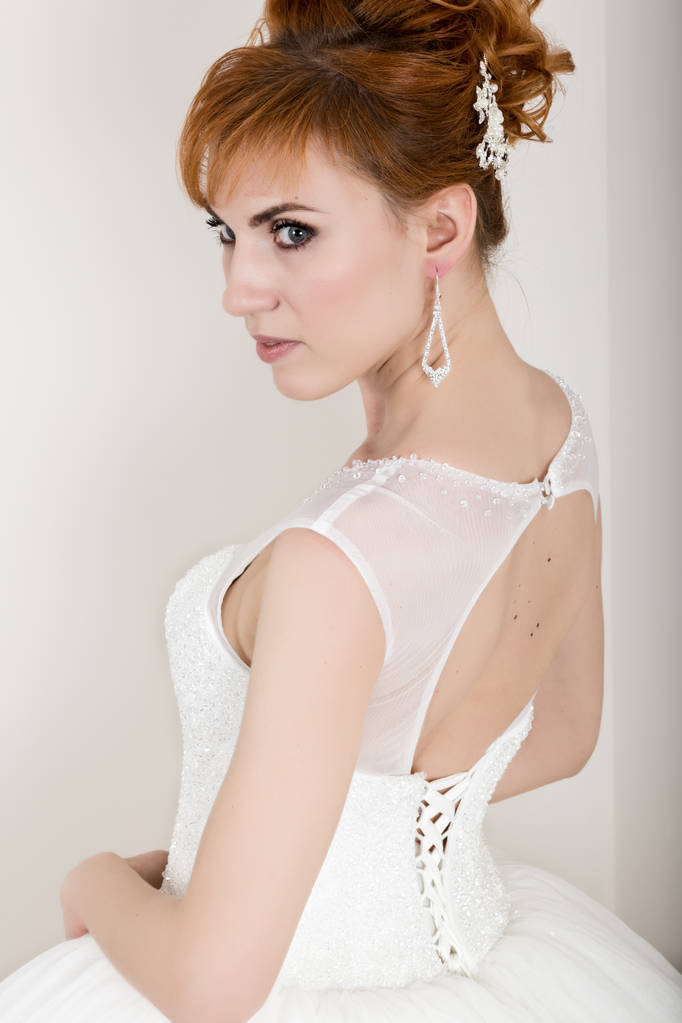 Όμορφη νεαρή κοκκινομάλλα νύφη φορώντας λευκό νυφικό με επαγγελματικό μακιγιάζ και χτένισμα - Φωτογραφία, εικόνα