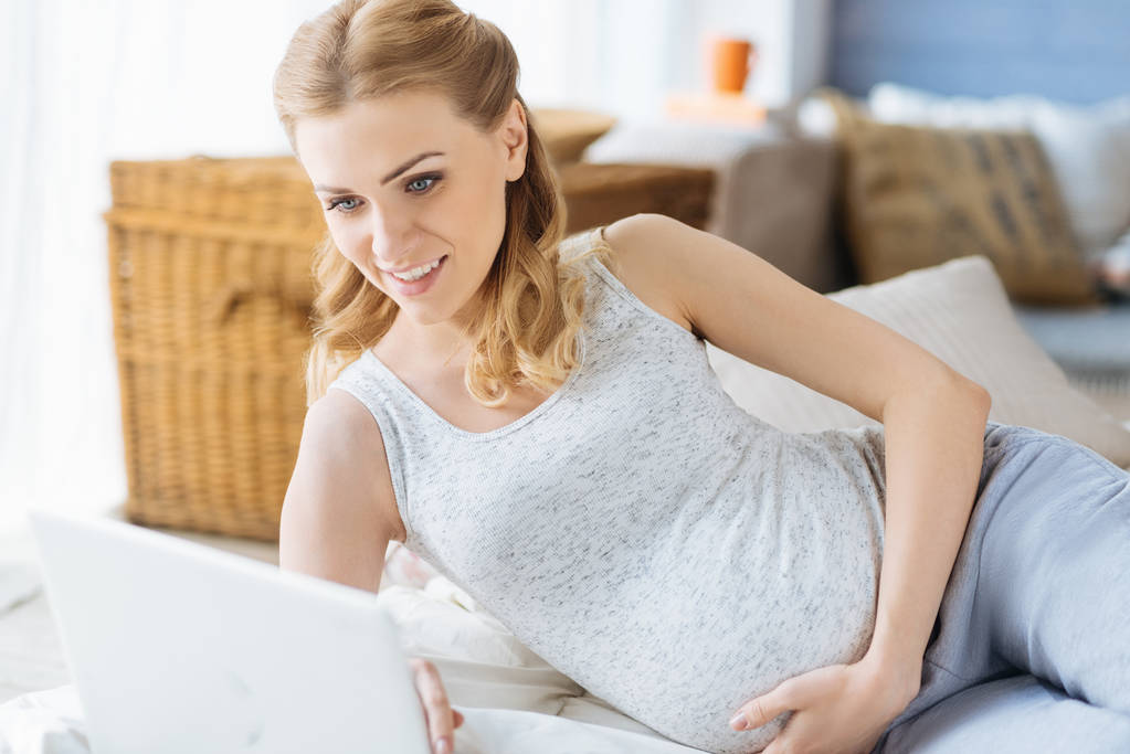 Iloinen raskaana oleva nainen käyttää kannettavaa makuuhuoneessa
 - Valokuva, kuva
