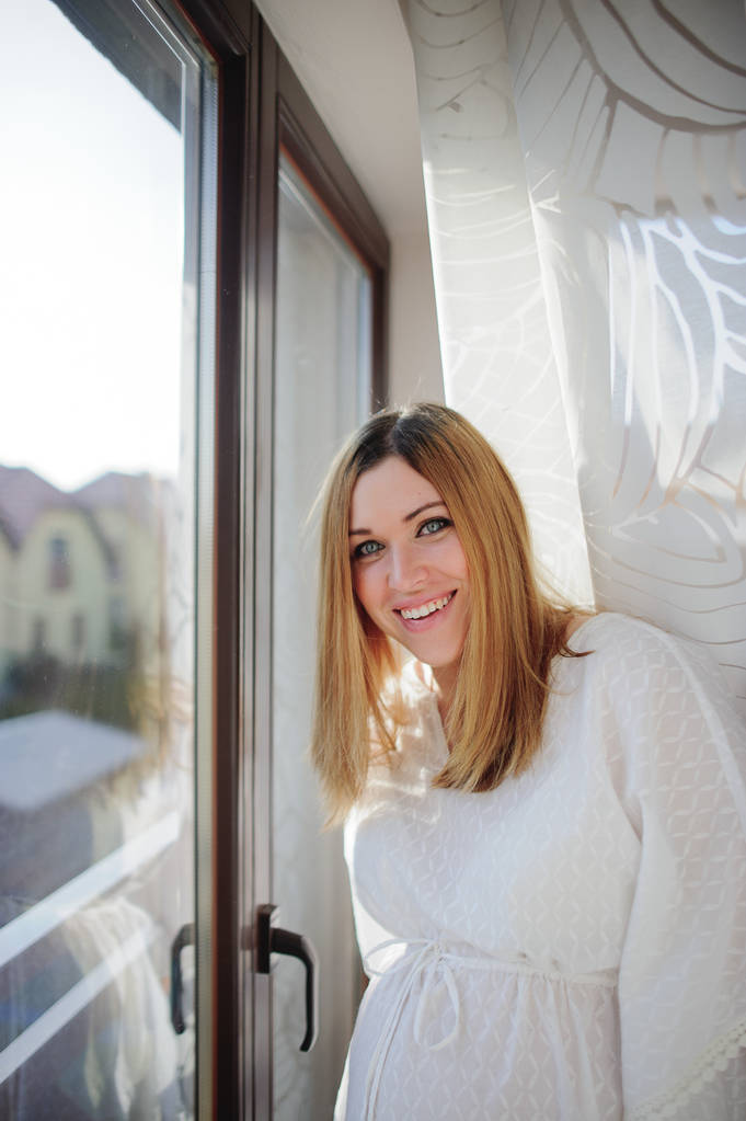 Γοητευτική έγκυος γυναίκα στέκεται κοντά σε ένα παράθυρο με λευκές κουρτίνες. - Φωτογραφία, εικόνα
