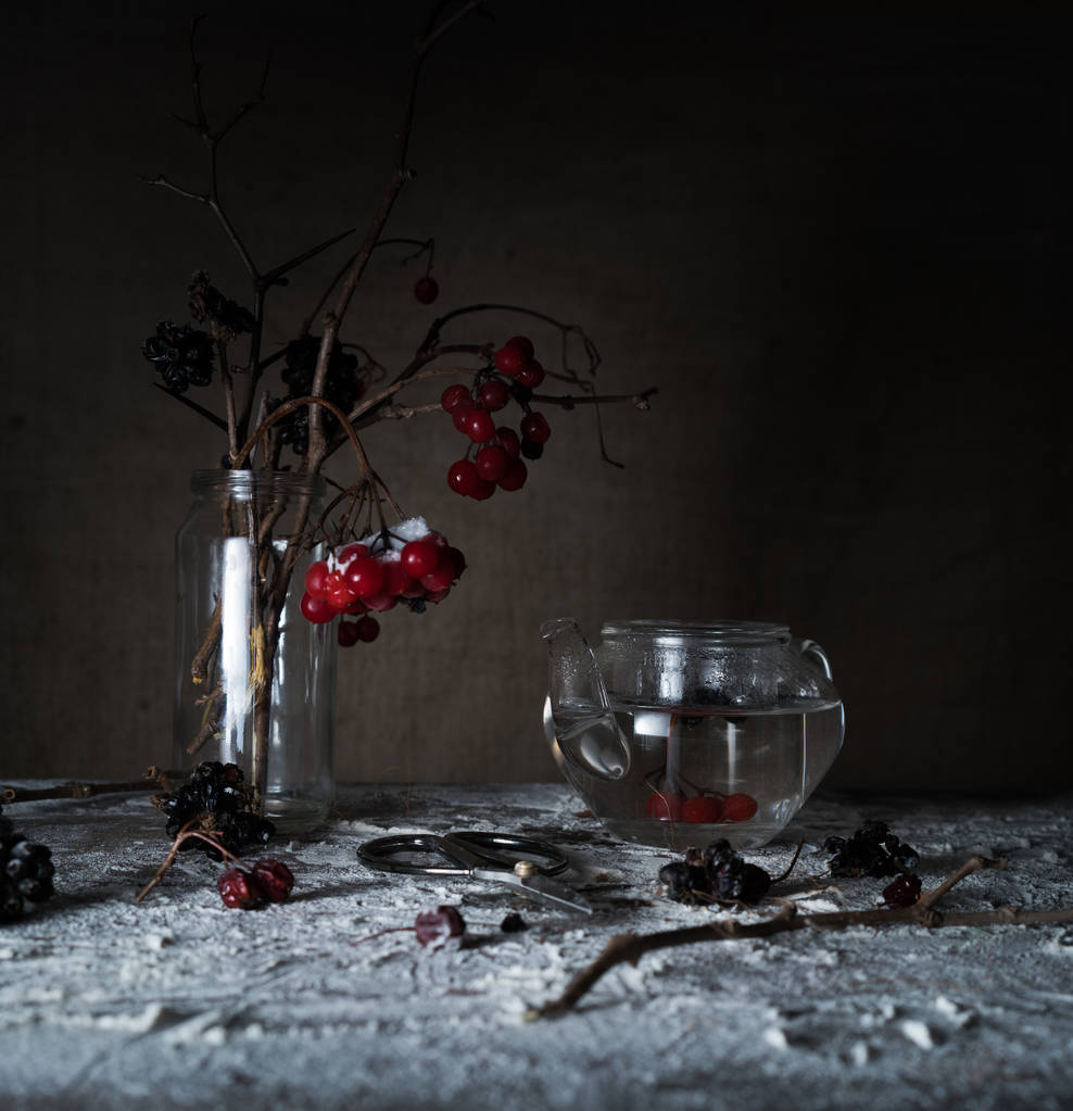 Stilleven. Viburnum takken met bessen en sneeuw in een doorzichtige vaas, glazen theepot op houten tafel - Foto, afbeelding