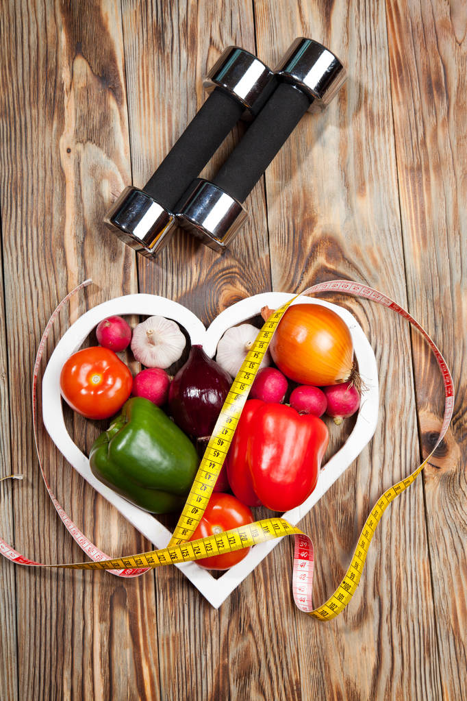 Sport i dieta. Warzywa, hantle centymetr. Papryka, pomidory, czosnek, cebula, rzodkiewki w serca na tle rustykalnym - Zdjęcie, obraz