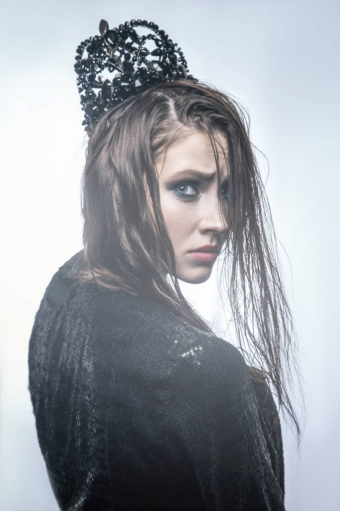 модный портрет молодой красивой девушки с макияжем и каблуками на лице с черной короной на голове и черным платьем. со светом сзади на светлом фоне
. - Фото, изображение