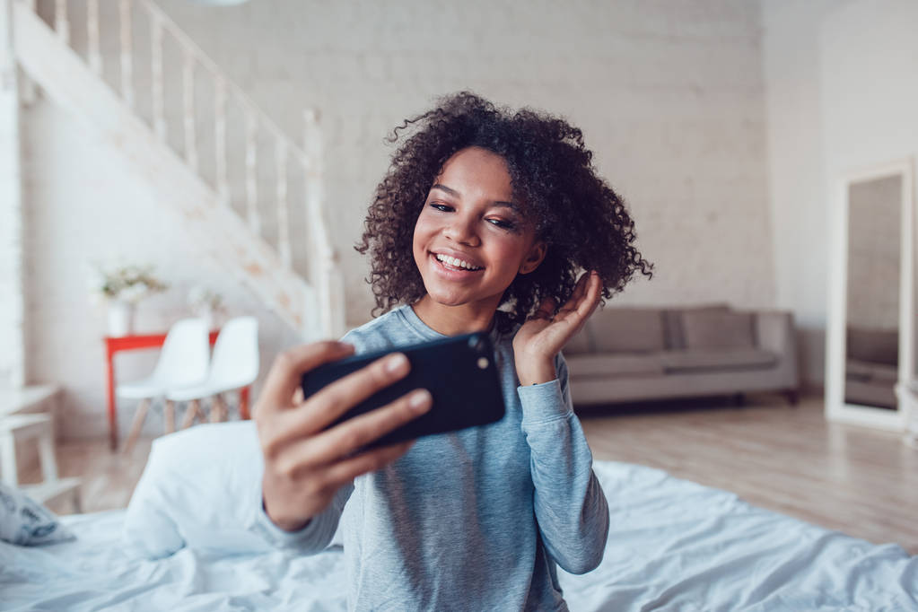 Pretty smiling african girl make selfie sitting on bed at home. - Photo, Image