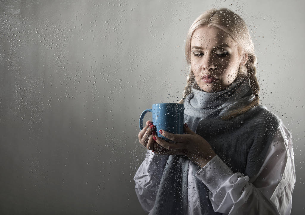 νεαρή γυναίκα, ντυμένη στο πουλόβερ πίνοντας καφέ ή τσάι, ποζάρει πίσω από διαφανές γυαλί που καλύπτεται από σταγόνες νερού - Φωτογραφία, εικόνα