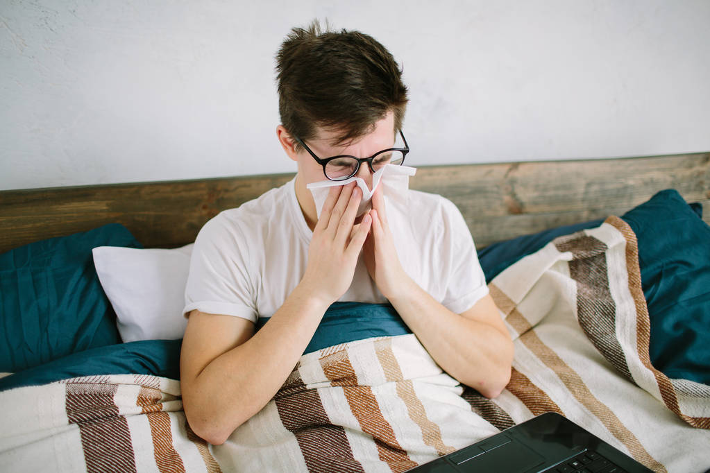 Egy fiatal férfi zsebkendővel. A beteg fickónak az ágyon folyik az orra. Az ember gyógymódot keres a megfázásra. - Fotó, kép