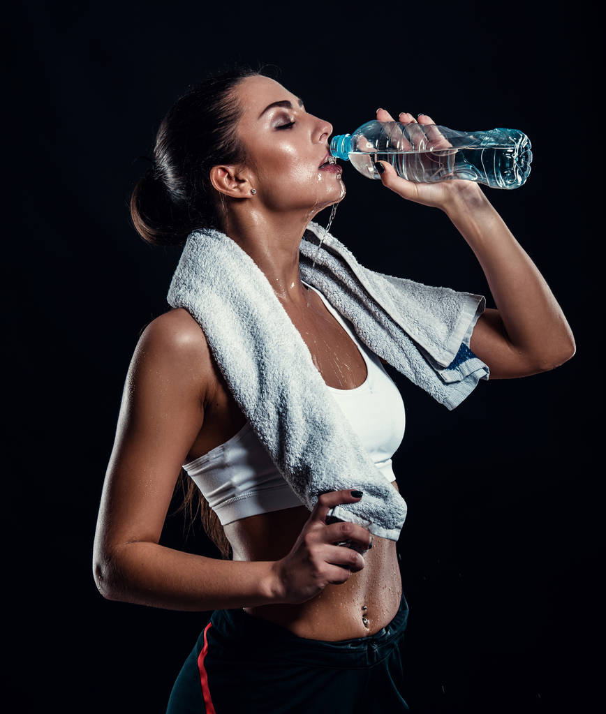 Привлекательная атлетичная молодая женщина с идеальным телом пьет воду из бутылки с полотенцем на шее на черном фоне. Фитнес-девушка, расслабляющаяся после тренировки
. - Фото, изображение