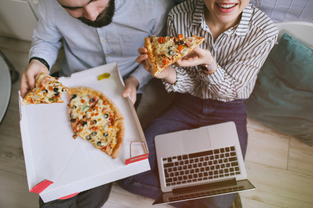 Jonge hipster mannelijke vrouwelijke paar thuis eten pizza met laptop - Foto, afbeelding