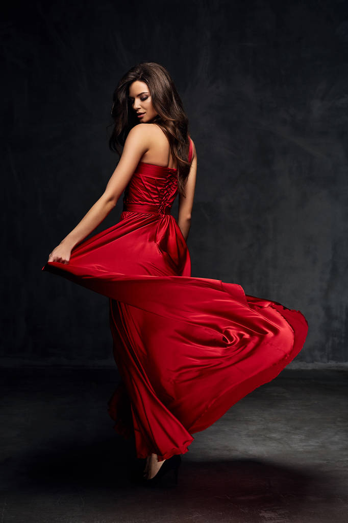 Όμορφη νεαρή σέξι μοντέλο θηλυκό με σκούρα μαλλιά στο εκπληκτικό μακρύ κόκκινο φόρεμα και μαύρα παπούτσια, ποζάρουν στο σκοτεινό θάλαμο - Φωτογραφία, εικόνα