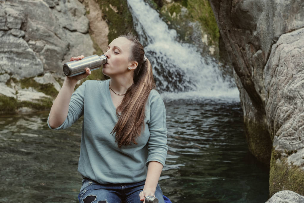 Молода жінка п'є воду з металевої пляшки на тлі вата
 - Фото, зображення