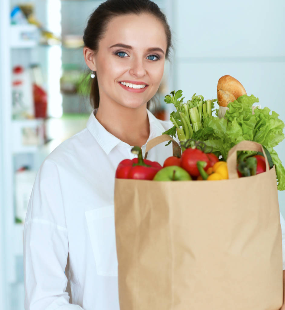 Fiatal nő gazdaság élelmiszer bevásárló táska zöldségekkel.Álló a konyhában - Fotó, kép
