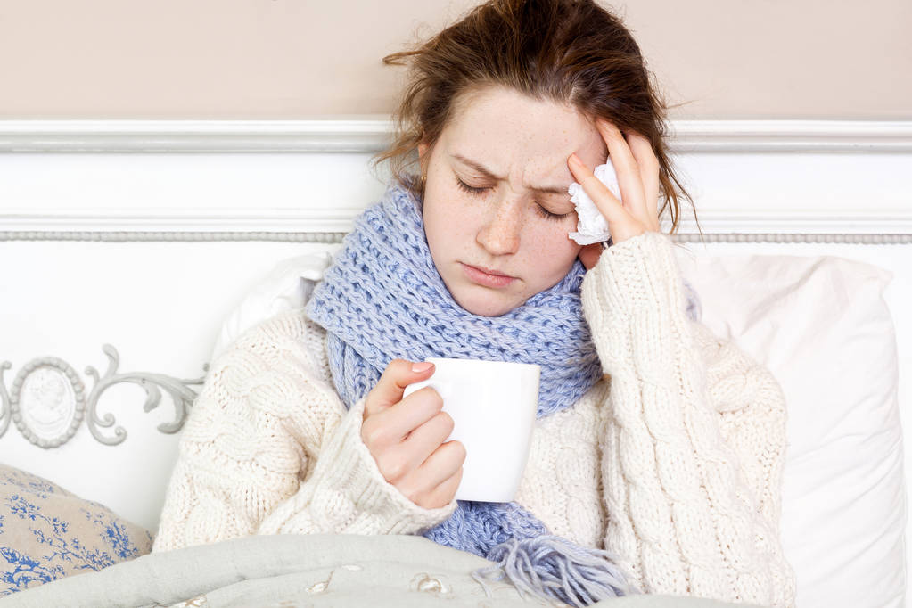 Sick woman with cup of tea. Closeup image of young frustrated woman in knitted blue scarf holding a cup of tea while sitting in bed of her room. - Photo, Image